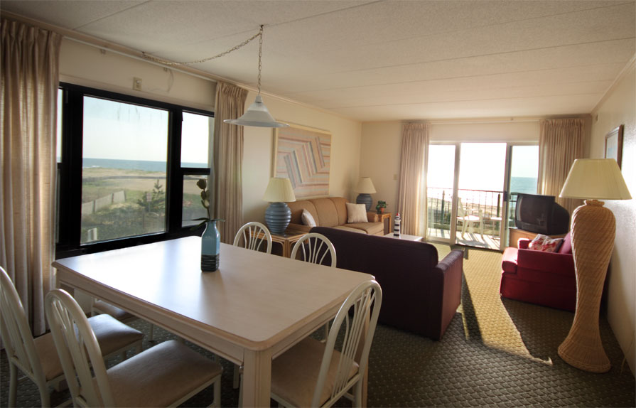 The living room and dining room are connected and inlcude a couch, love seat, arm chair, tv, table with chairs and large windows with views of the ocean.